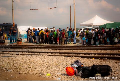 A group of people waiting at a border surveilled by border guards